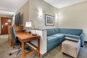 A hotel room with a wooden desk, a blue sofa, a small ottoman, a flat-screen TV, and a framed picture on the wall. - Drury Plaza Hotel Phoenix Tempe (Tempe)