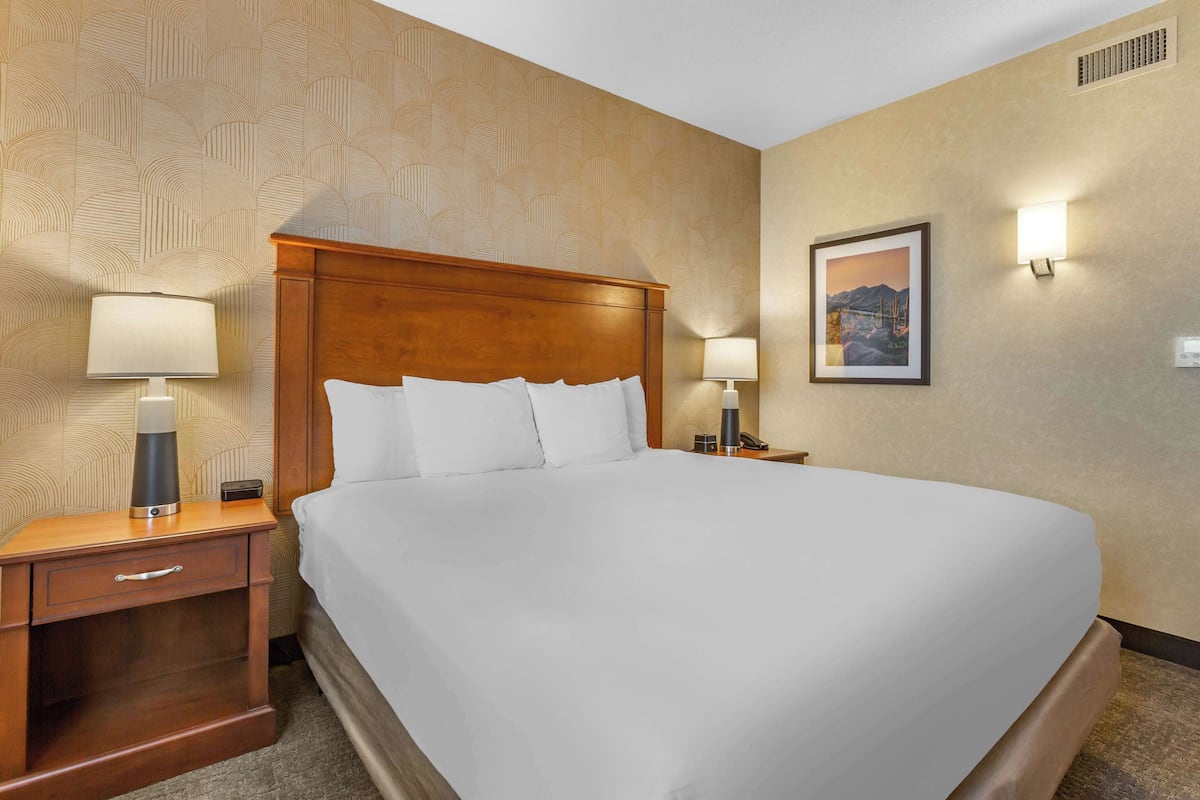 A neatly made bed in a hotel room with a wooden headboard, a nightstand with a lamp, and a wall-mounted picture.