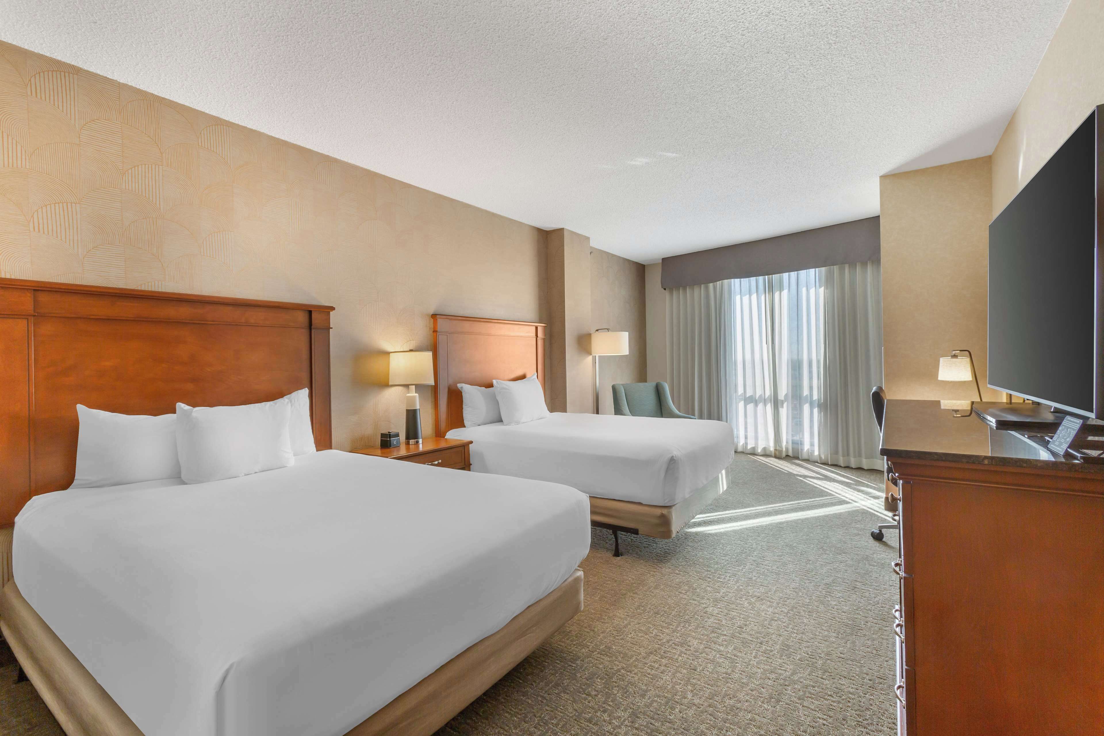 A hotel room with two beds, a desk, a TV, and a window with curtains.