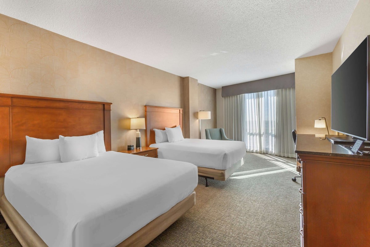 A hotel room with two beds, a desk, a TV, and a window with curtains.