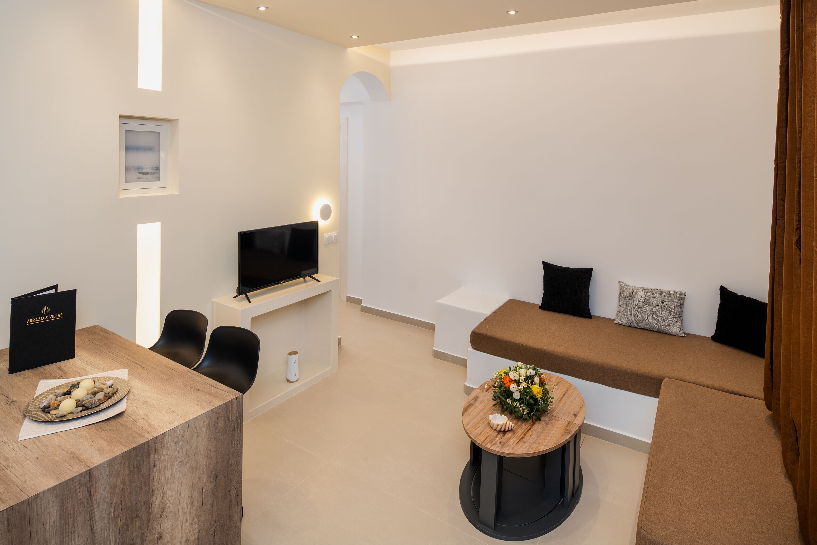 Uma sala de estar moderna com um sofá, uma TV, uma mesa de centro com flores e uma área de jantar com mesa e cadeiras.