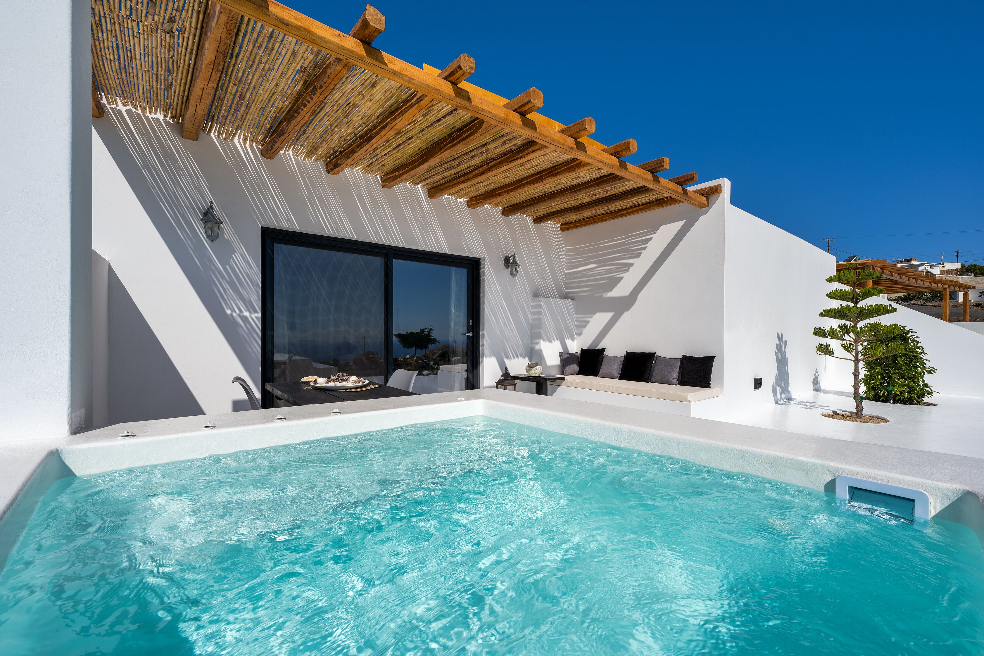 A modern outdoor pool area with a wooden pergola, a seating area with cushions, and a glass door leading to a covered space.