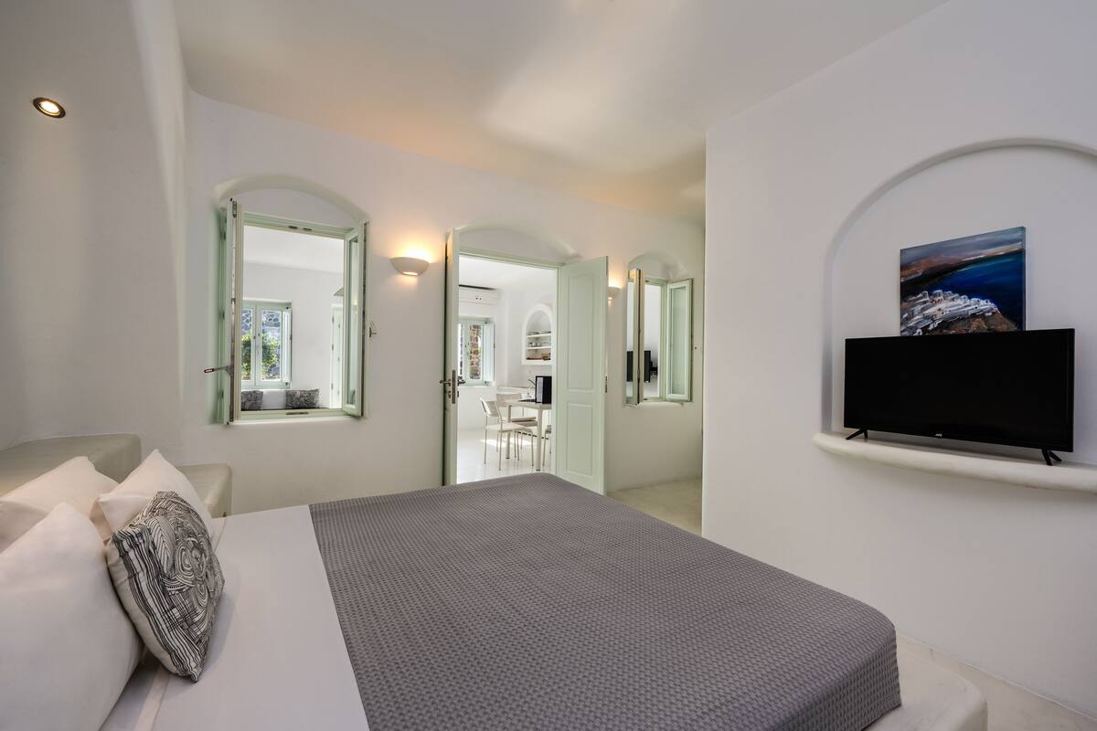 A modern hotel room with a large bed, a flat-screen TV mounted on the wall, and a view of the sea through the window.