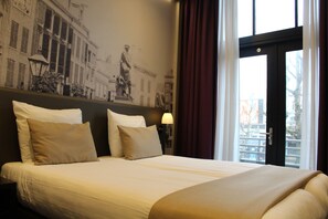 A hotel room with a large bed, a headboard with a mural, a nightstand with a lamp, and a window with curtains.