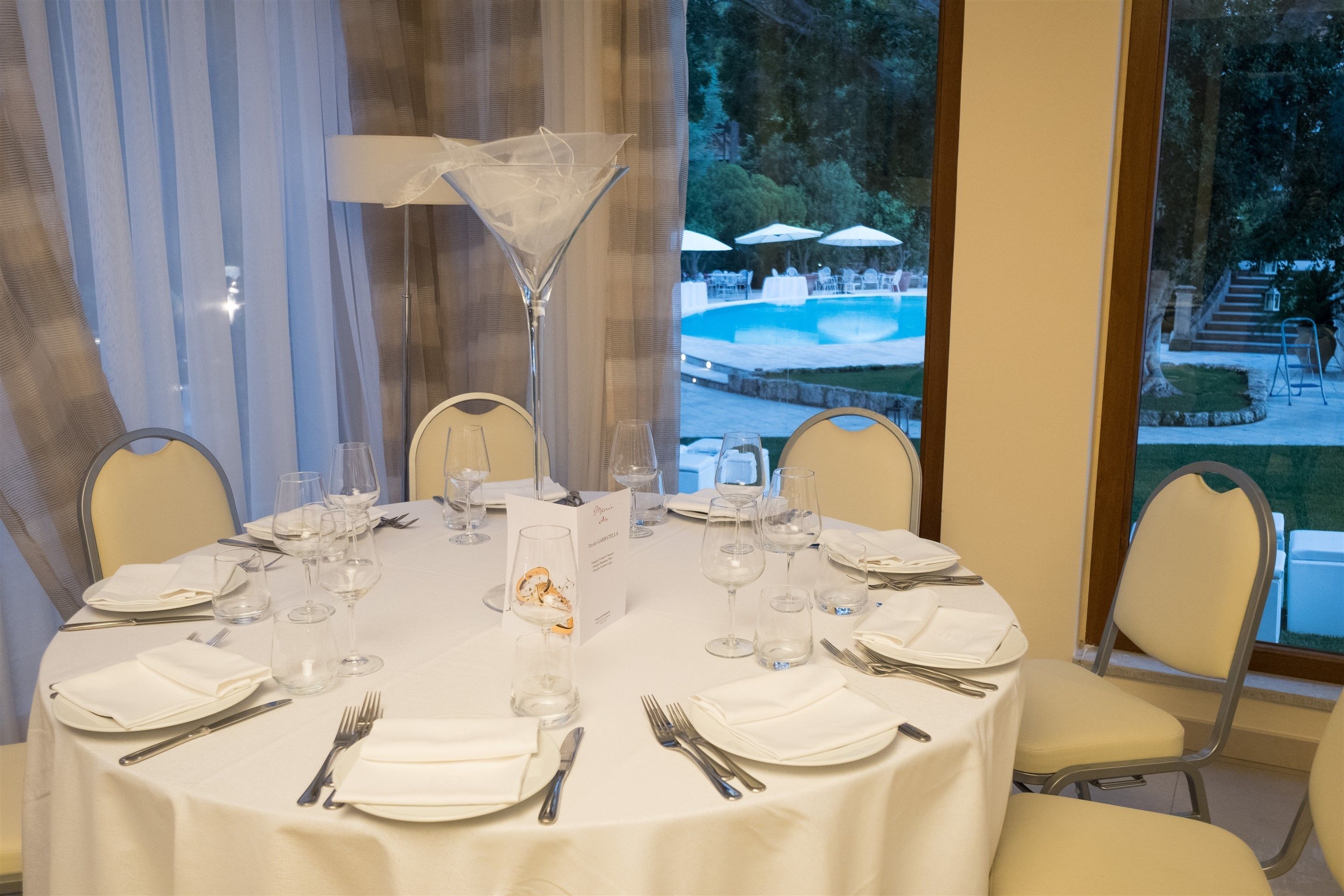 A round table set for six with white tablecloths, clear glassware, and metal cutlery.