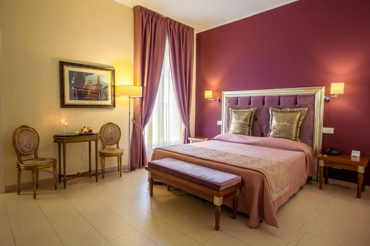A hotel room with a large bed, a bench, two chairs, a small table, and a painting on the wall.