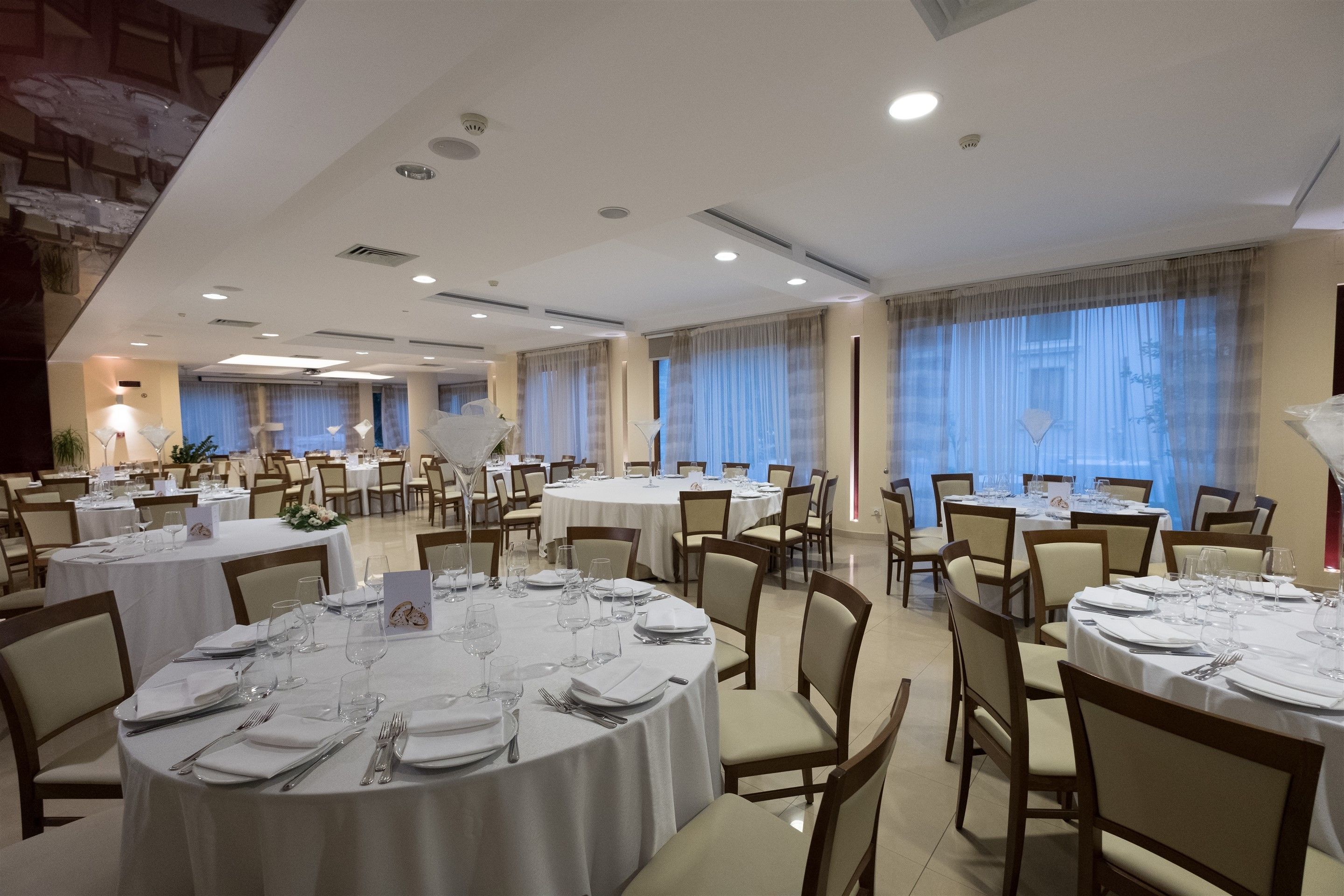 A spacious banquet hall with round tables set for dining, featuring a high ceiling with recessed lighting and large windows with curtains.