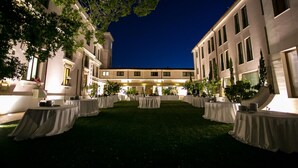 A courtyard with tables set up for an event, surrounded by buildings with large windows and a well-lit exterior. - Hotel Parco delle Fontane (Syracuse)