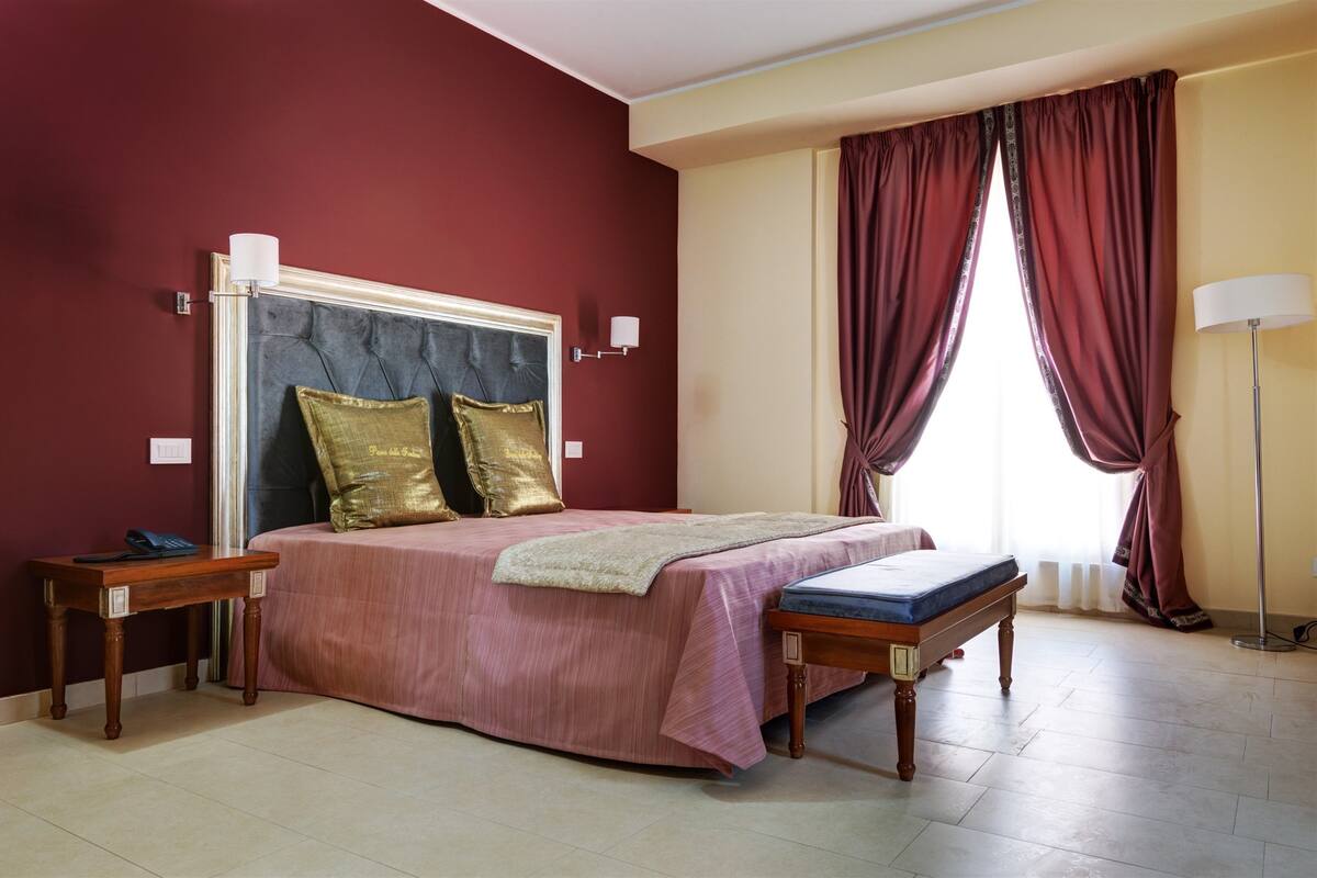 A bedroom with a bed, two nightstands, a bench, a lamp, and a window with curtains.
