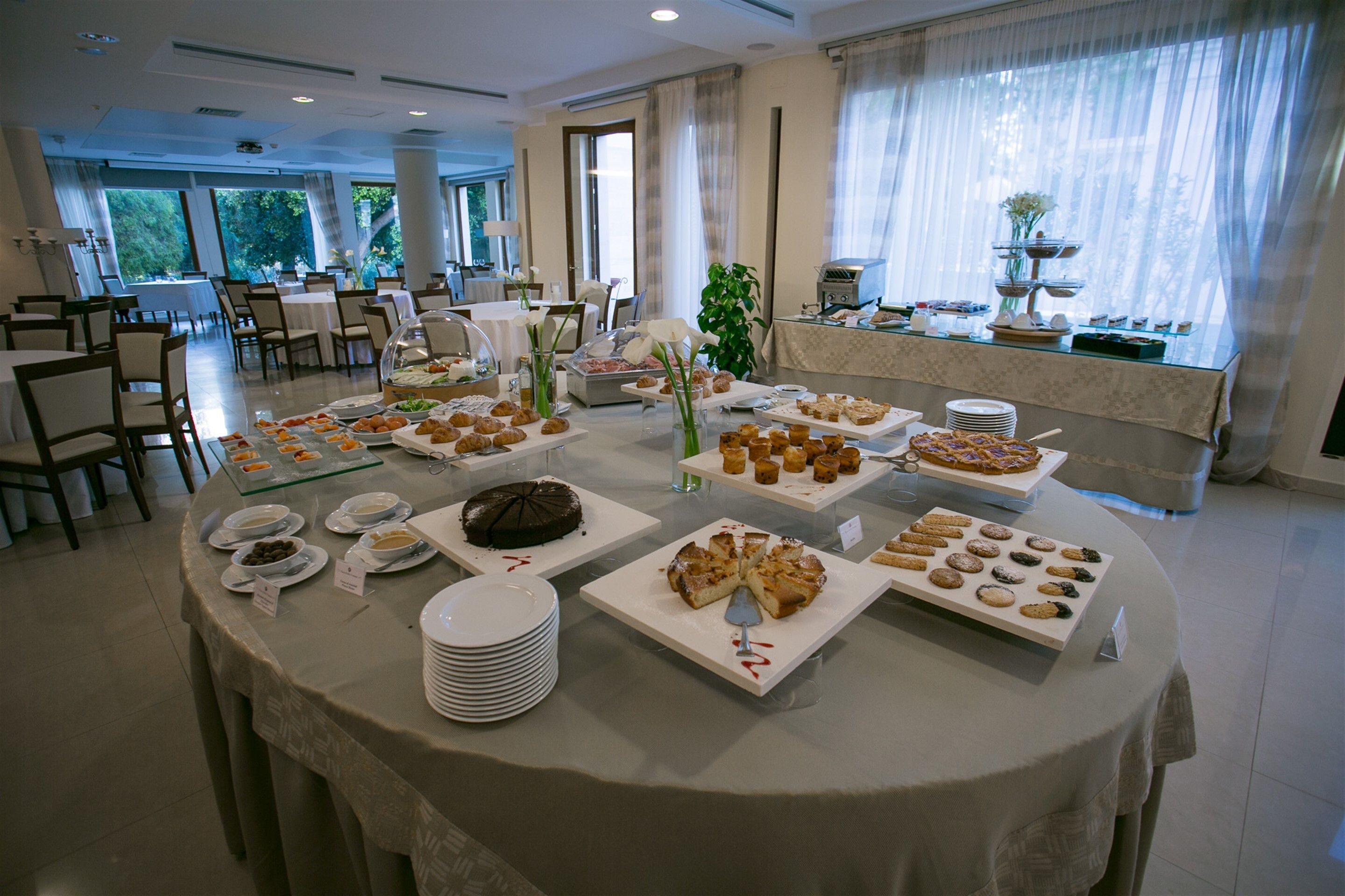 A buffet setup with various food items and a cake on a round table in a well-lit room with large windows and a view of greenery outside.
