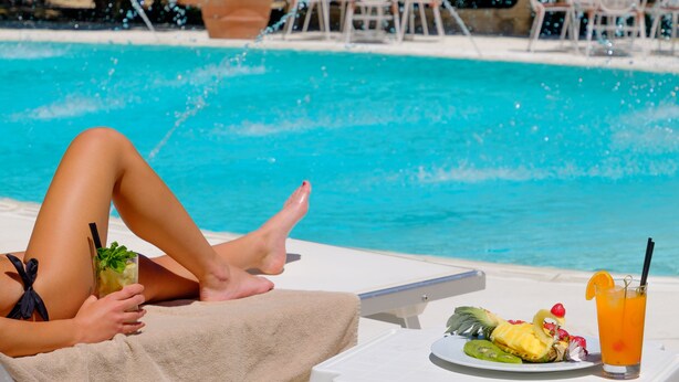 A poolside setting with a person relaxing, a table with a drink and fruit, and a view of the pool and chairs in the background. - Hotel Parco delle Fontane (Syracuse)