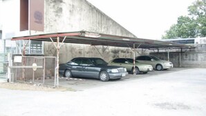 A parking area with a covered structure, two cars parked under it, and a building in the background. - SB Apartment (Bangkok)