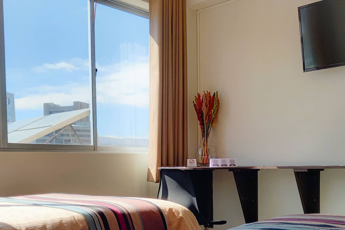 A hotel room with two beds, a desk with a computer, a TV on the wall, and a window with a view of buildings.