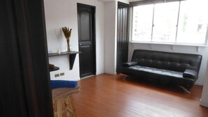 A living room with a black leather sofa, wooden floor, a small stool, a shelf with a vase, and a door. - Hotel Sagarnaga (La Paz)