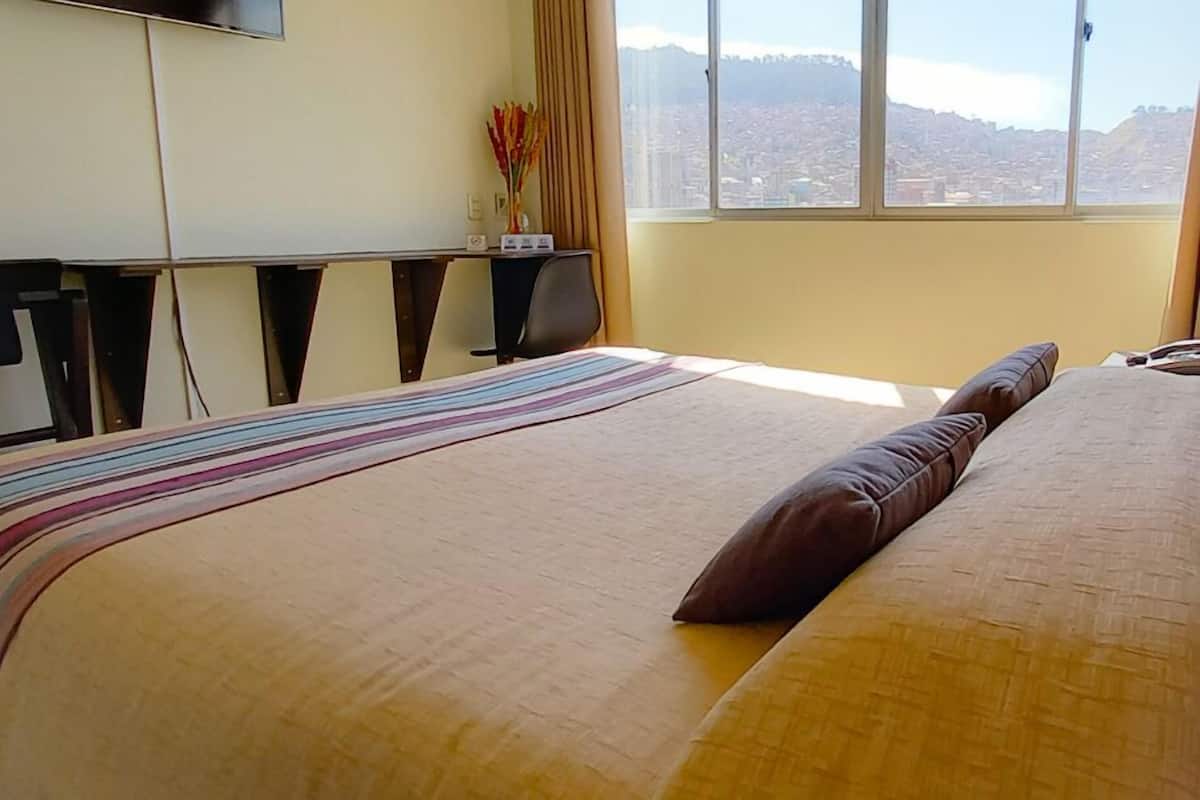 A hotel room with a large bed, a desk, and a window with a city view.