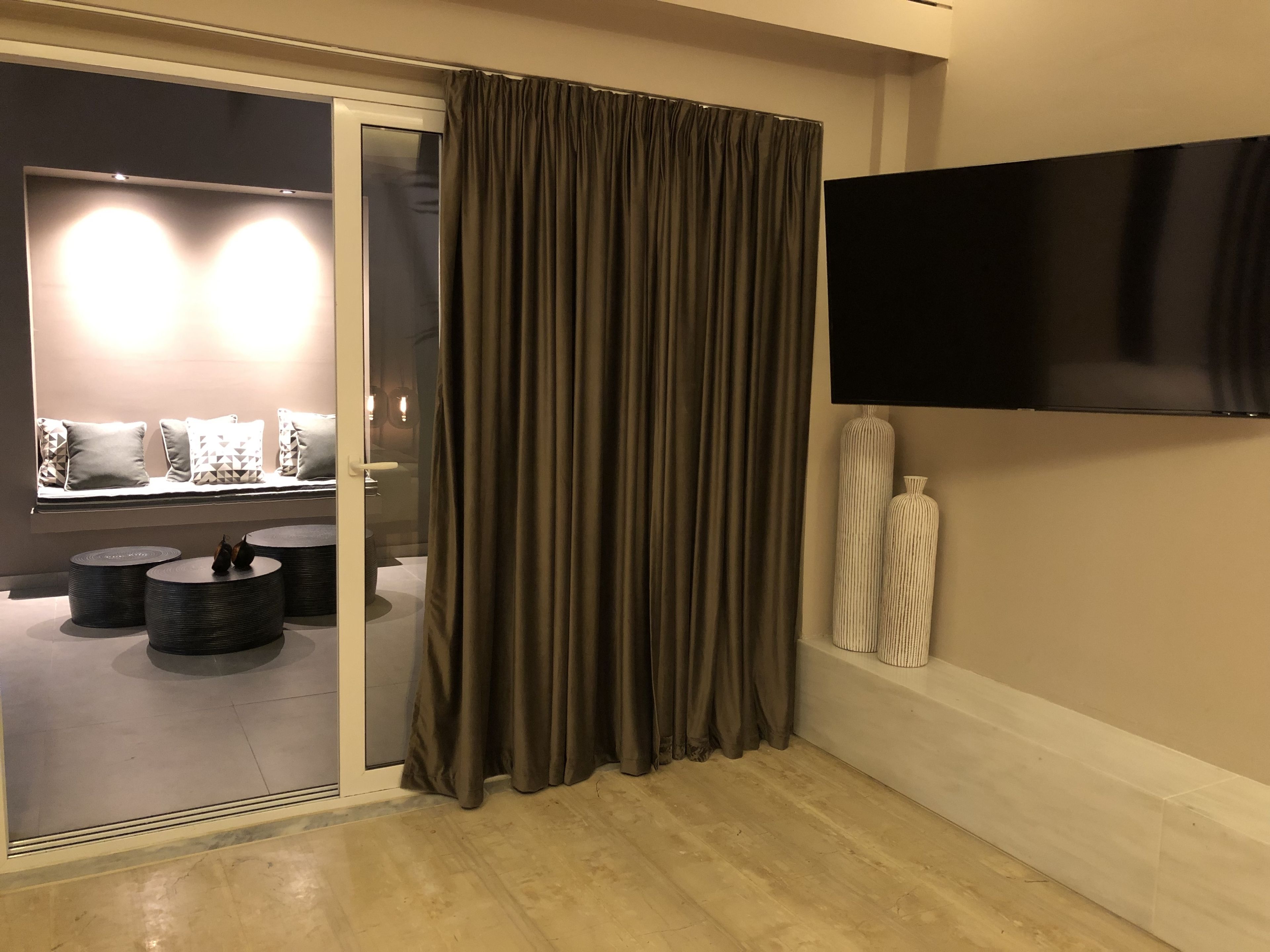 A modern living room with a sliding glass door leading to a balcony, a flat-screen TV mounted on the wall, and a seating area with cushions and a coffee table.