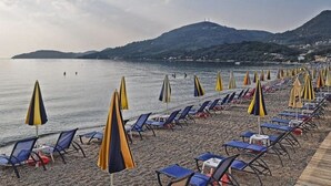 A beach with rows of sun loungers and umbrellas, a calm sea, and a mountain in the background.