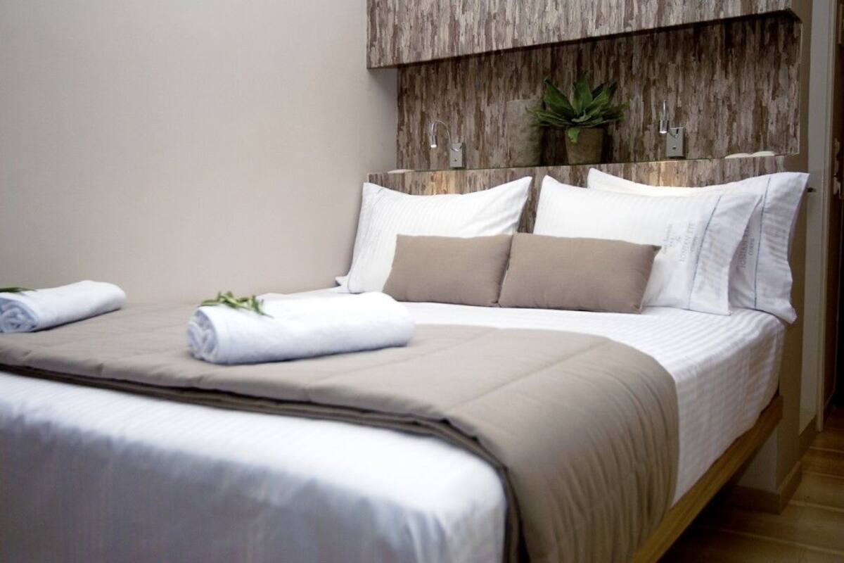 A neatly made bed with white and beige bedding, a wooden headboard, and a small potted plant on the wall.