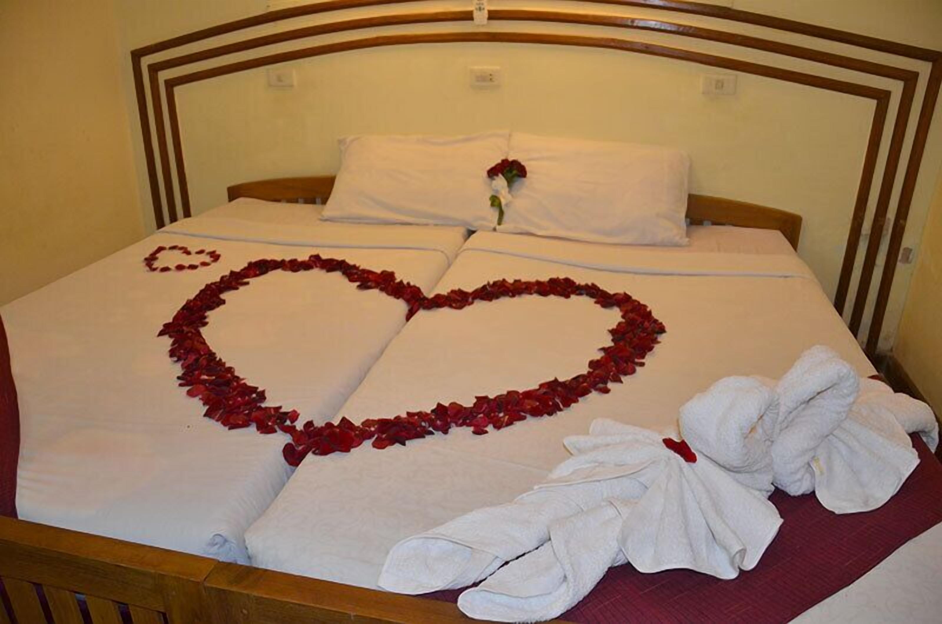 A neatly made bed with rose petals forming a heart shape on the bedspread and a pair of towels folded on the side.