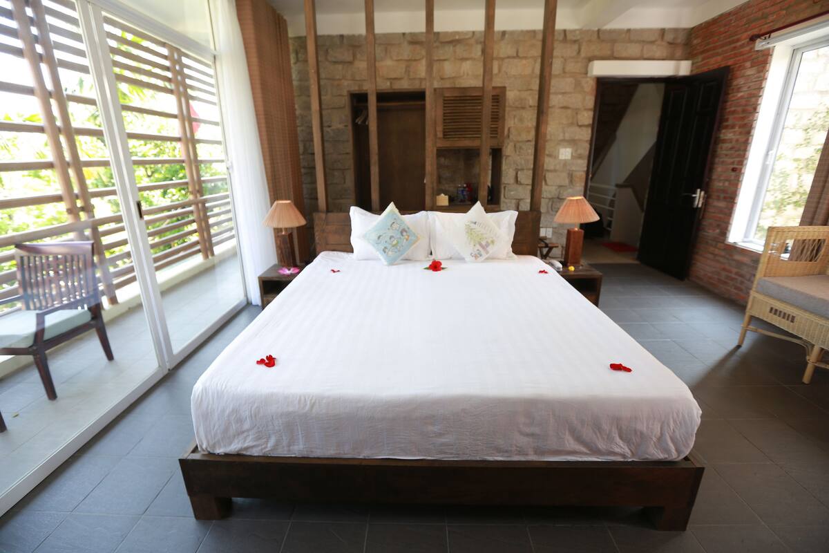 A bedroom with a large bed, wooden headboard, and a stone wall. There is a window with blinds and a chair by the window.