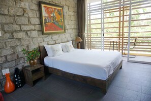 A bedroom with a stone wall, a bed with white bedding, a small nightstand with a plant, and a window with a view of greenery.