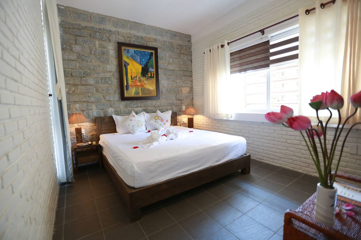 A hotel room with a large bed, a stone wall, a painting, and a window with curtains.