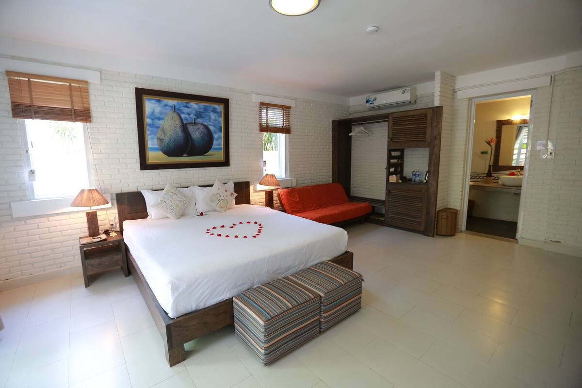 A bedroom with a large bed, a red sofa, a bench, a painting, and a bathroom visible through an open door.