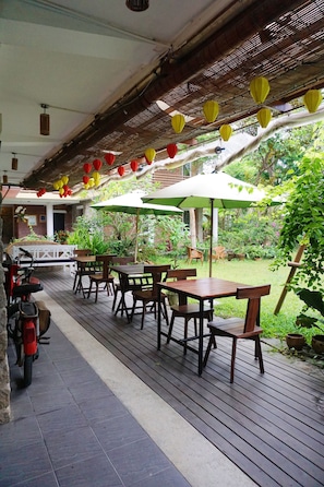 An outdoor patio with wooden tables and chairs, surrounded by greenery and a bamboo ceiling.