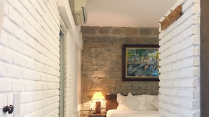 A bedroom with a white brick wall, a stone accent wall, a bed with white linens, a nightstand with a lamp, and a framed picture on the wall. - Rock Villa Hoi An (Da Nang)