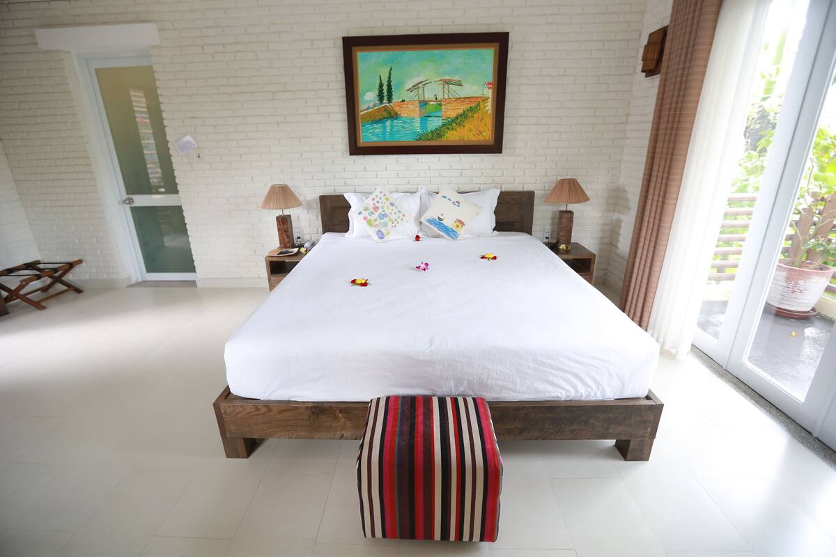 A bedroom with a large bed, a wooden bench, a painting on the wall, and a view of the outdoors.
