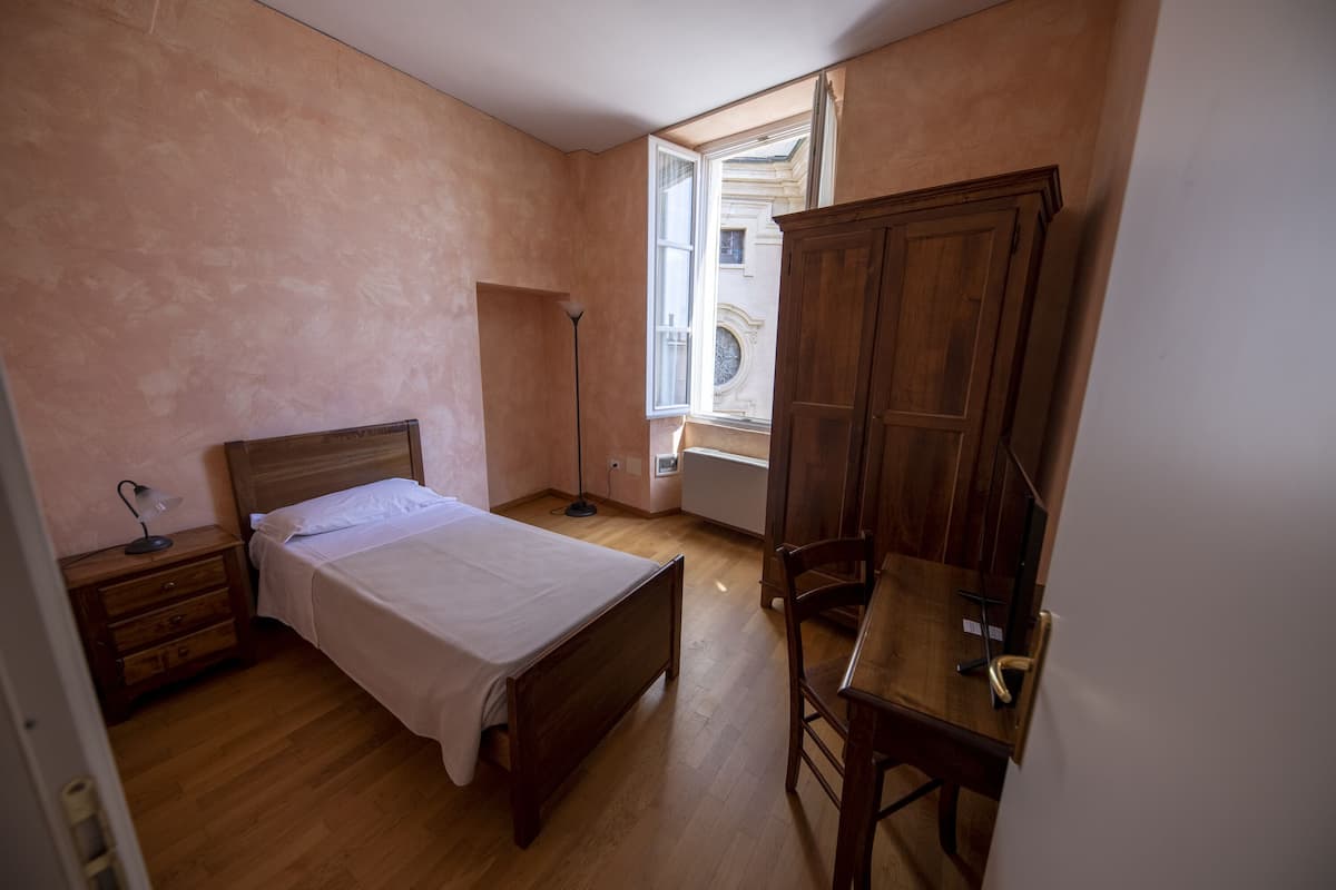 A bedroom with a wooden bed, a desk, a chair, a floor lamp, and a window with an open shutter.