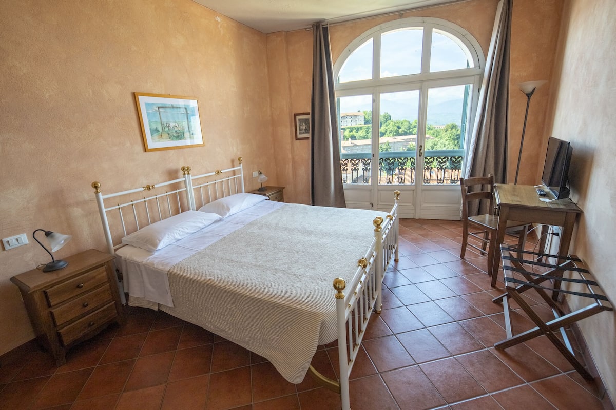 A bedroom with a bed, a nightstand, a desk, a chair, a window with a view, and a painting on the wall.