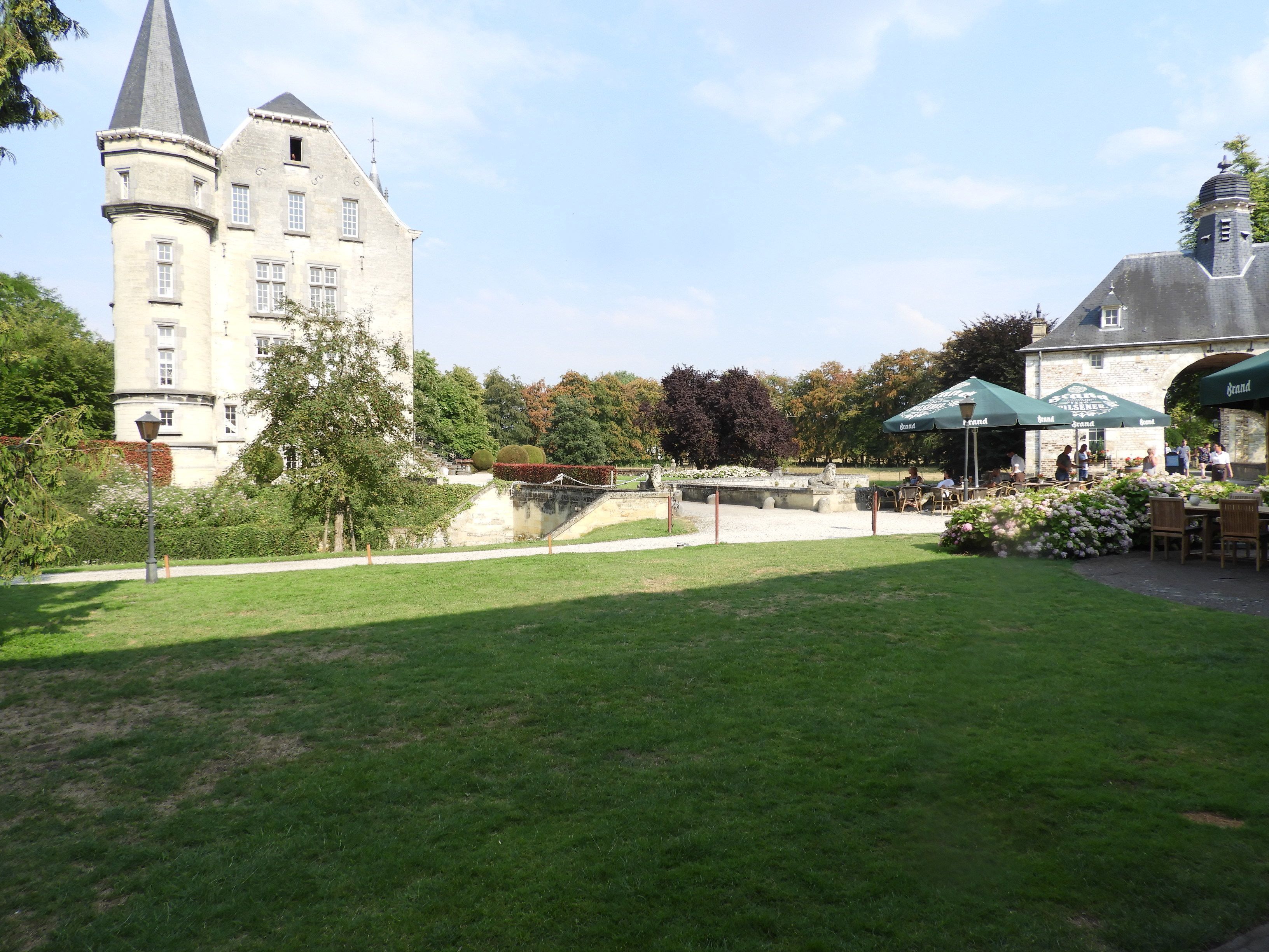 Eine Burg mit einem Turm, ein Garten mit Teich und Sitzgelegenheiten im Freien unter Sonnenschirmen.