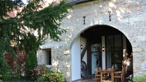 A stone building with a large arched entrance, a wooden table and chairs outside, and a wheel visible inside. - Kasteel Schaloen (Schin op Geul)