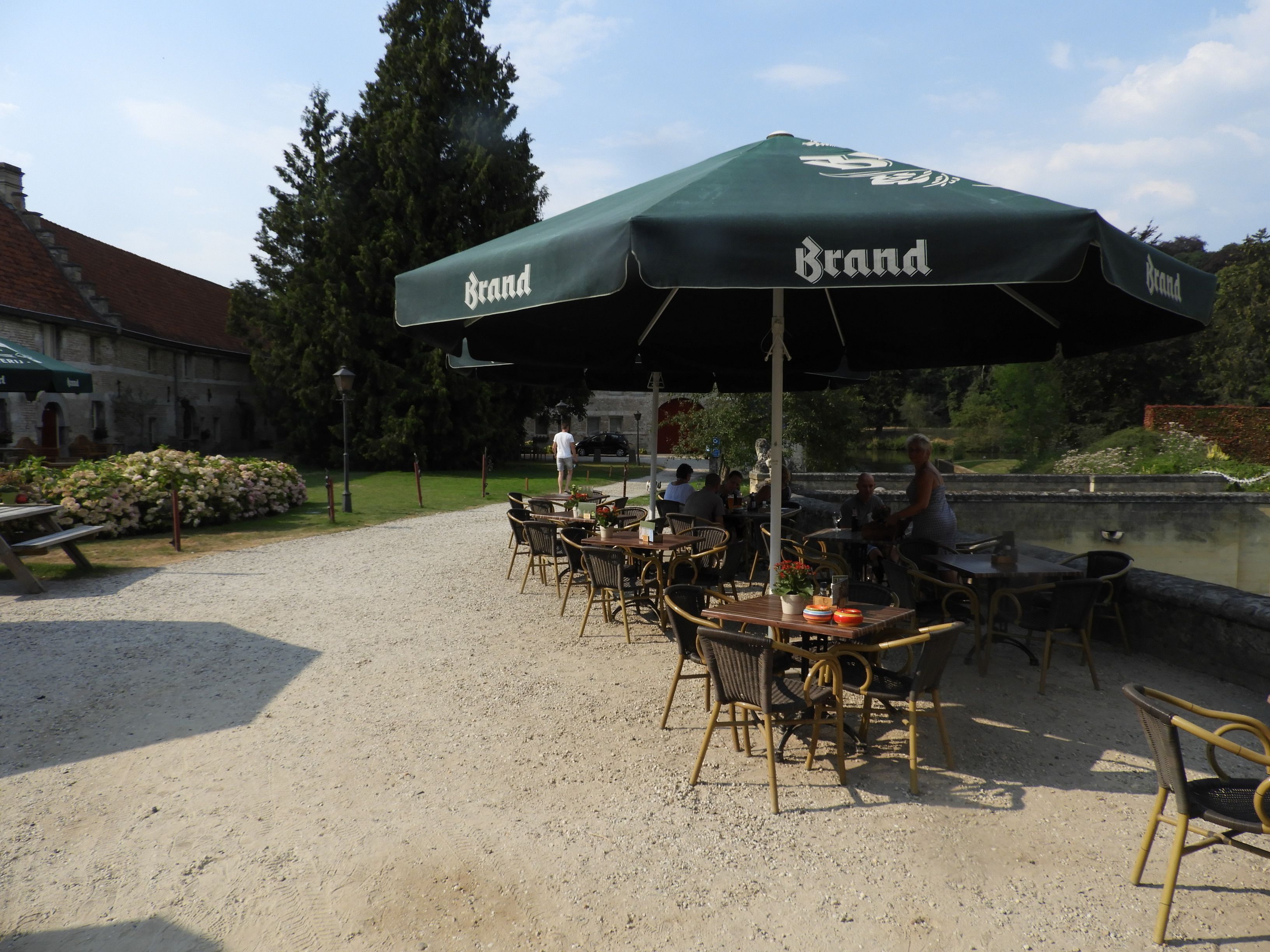An outdoor cafe with tables and chairs under green umbrellas, a gravel path, and a building in the background.