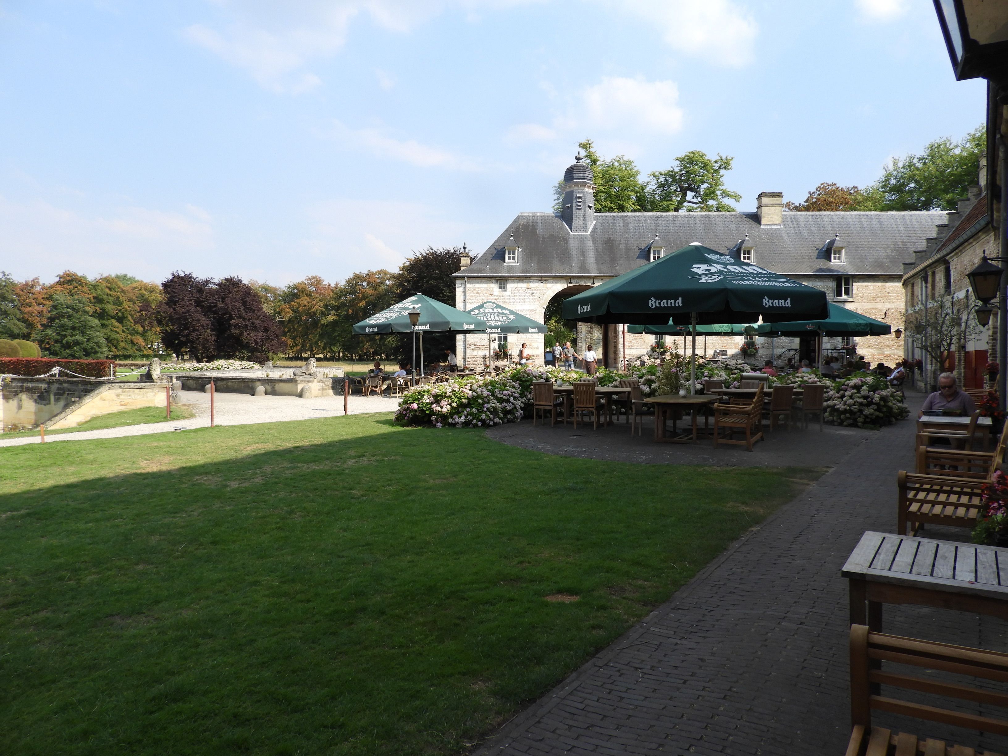 Ein Straßencafé mit Tischen und Stühlen unter grünen Sonnenschirmen, im Hintergrund ein Steinhaus und eine Rasenfläche.