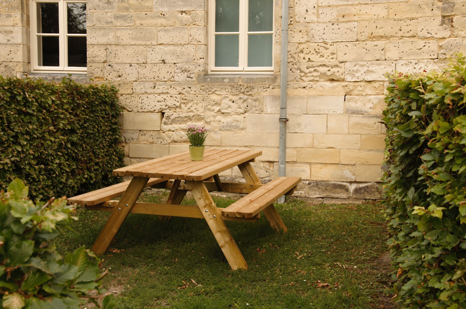 A wooden picnic table with benches in a garden area with stone walls and a hedge.