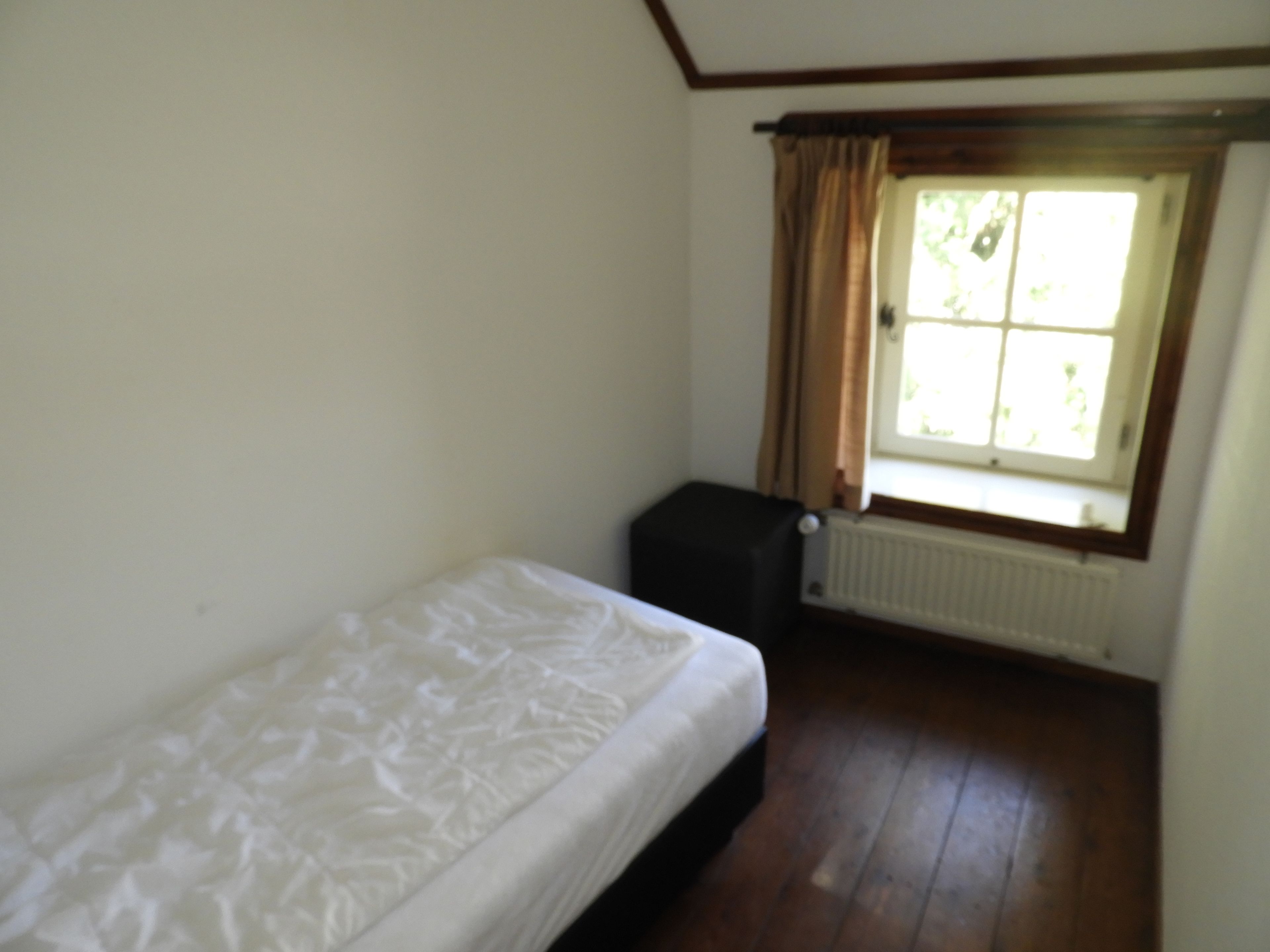 Une petite pièce, peu meublée, avec un lit simple, un radiateur et une fenêtre avec des rideaux.