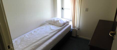 A small, single-bed room with a window, a radiator, and a wooden dresser.