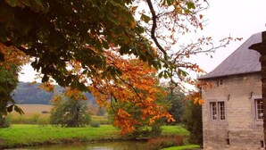 A stone building with a sloped roof, situated beside a calm water body, surrounded by greenery and trees with autumn foliage. - Kasteel Schaloen (Schin op Geul)