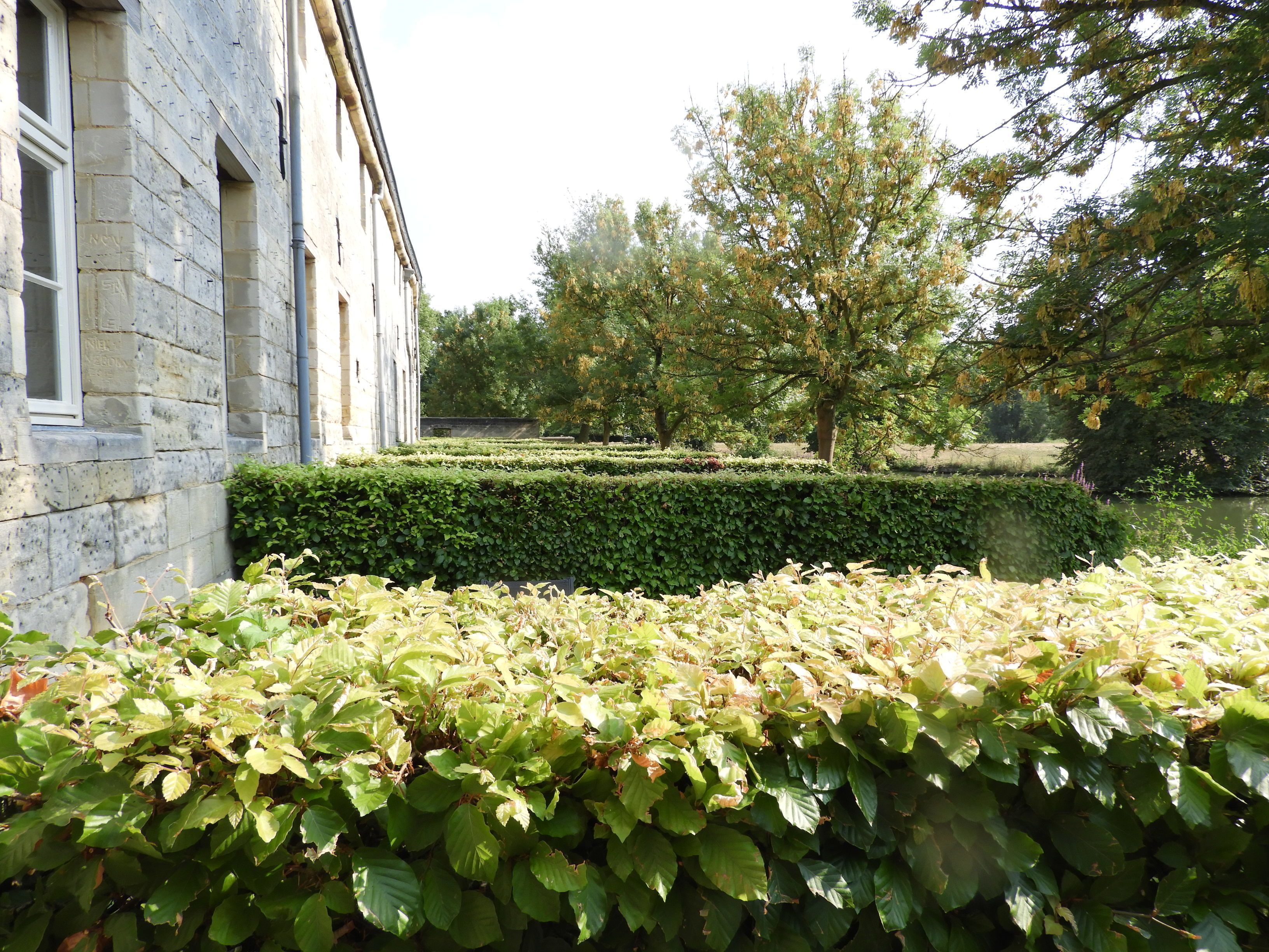 A stone building with a hedge in the foreground, a well-maintained garden, and trees in the background.