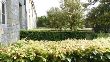 A stone building with a hedge in the foreground, a well-maintained garden, and trees in the background.