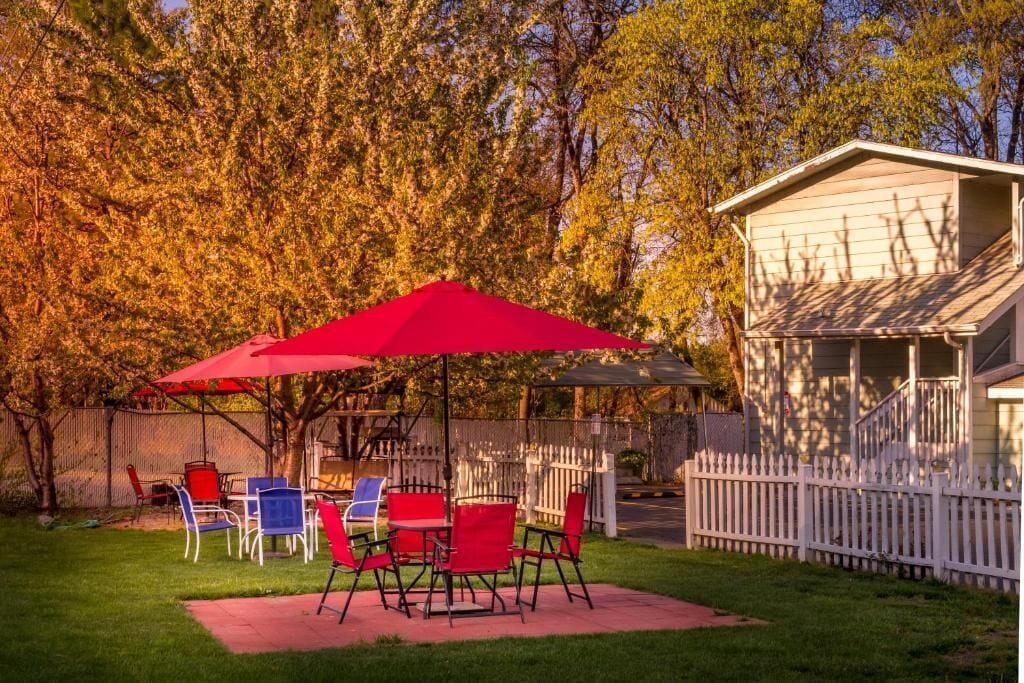 Une terrasse avec des chaises rouges et des parasols, une clôture blanche en bois et des arbres en arrière-plan.