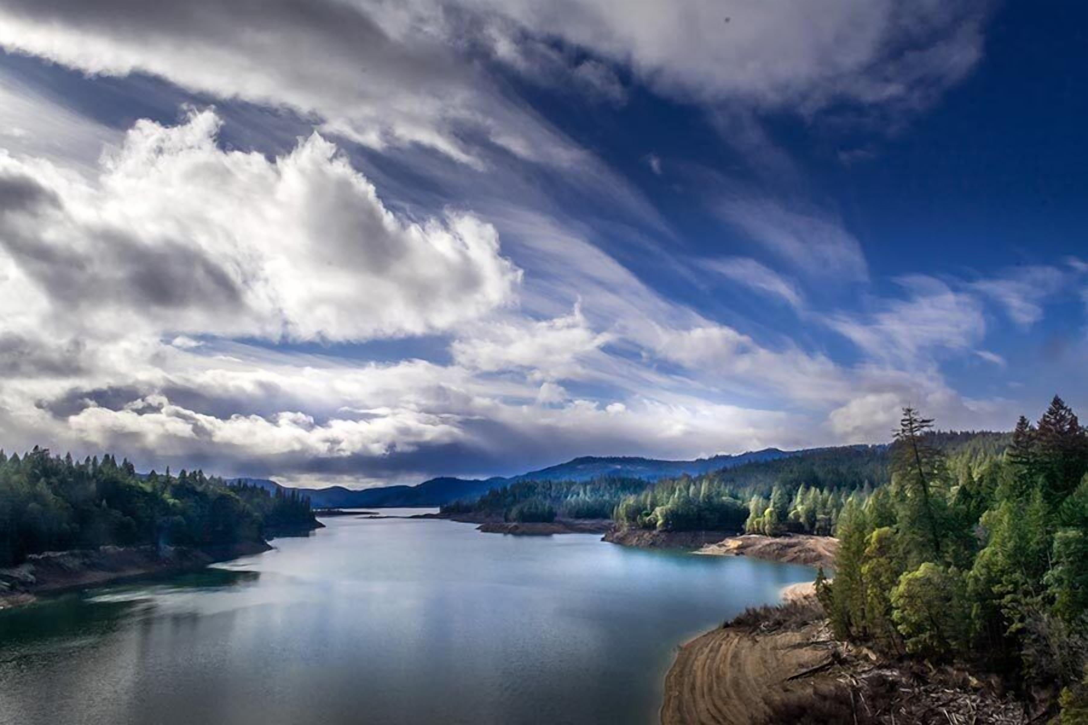 A serene landscape with a calm lake, dense forest, and distant mountains under a partly cloudy sky.