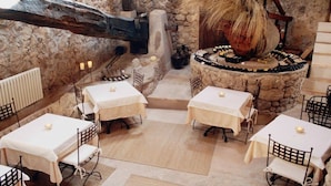 A rustic dining area with stone walls, wooden beams, and a central stone table surrounded by chairs. Tables are covered with white tablecloths and candles are placed on them. - Hotel Mirabó De Valldemossa (Valldemossa)