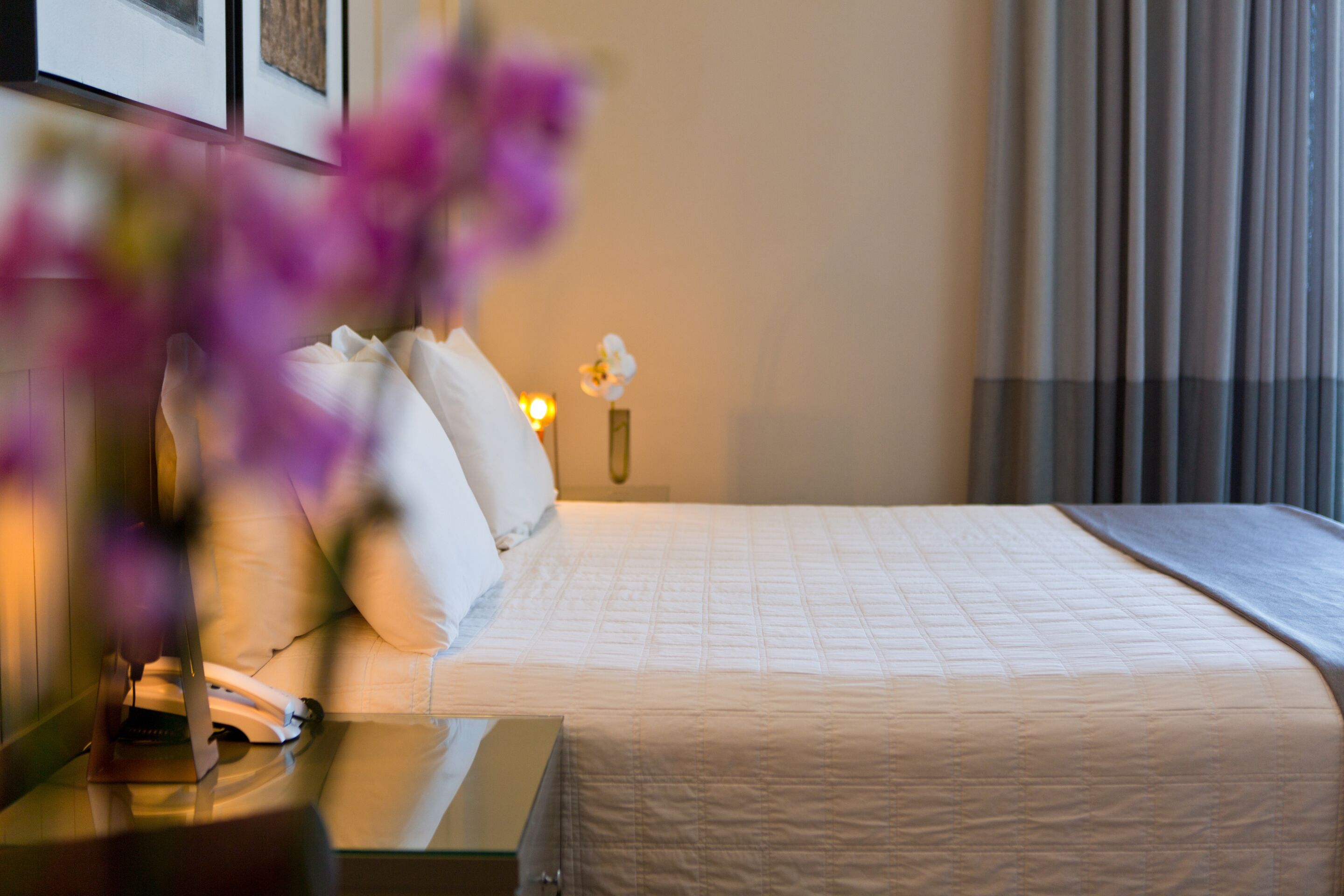 A hotel room with a bed, a bedside table with a lamp, and a vase with flowers.