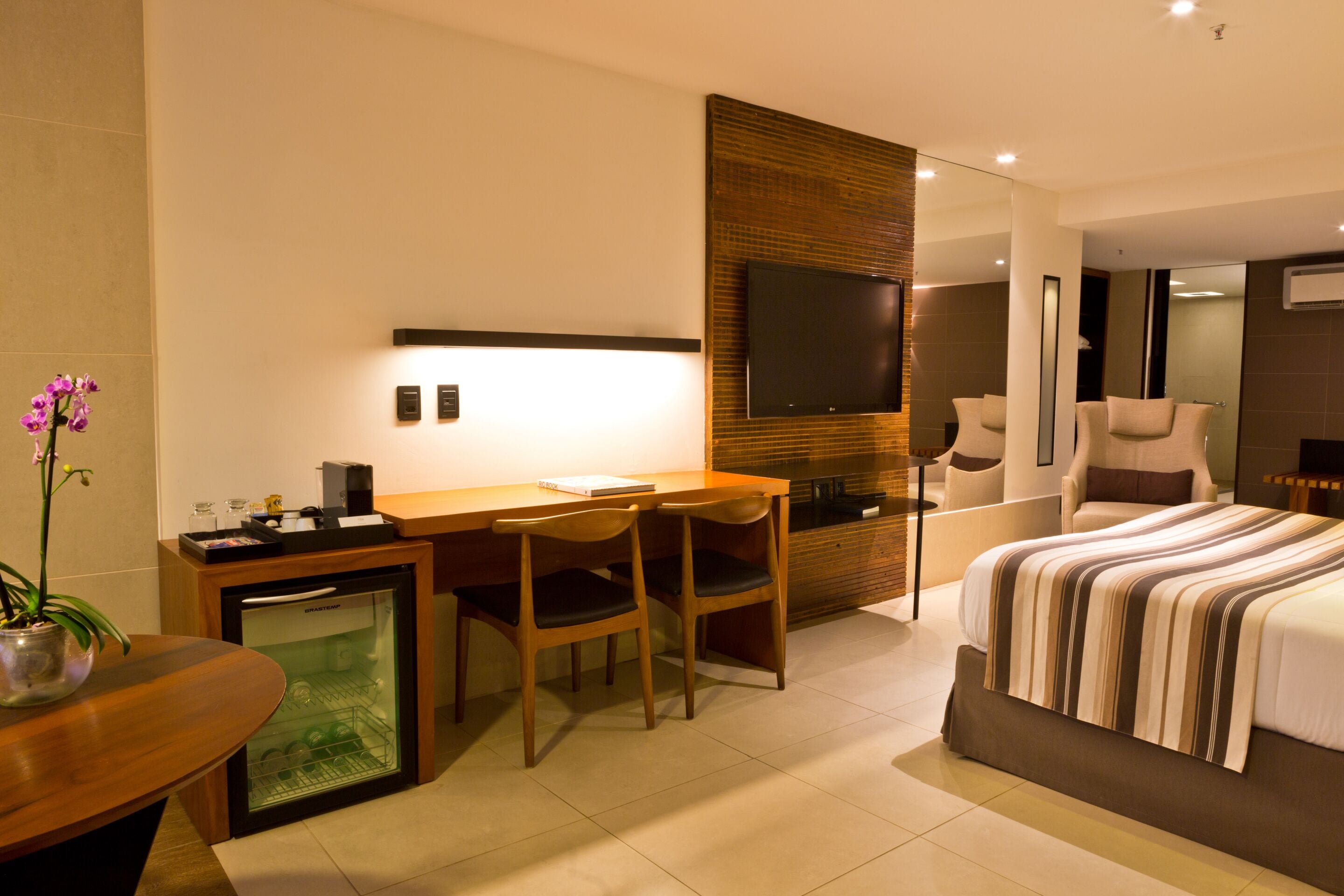 Une chambre d’hôtel moderne équipée d’un lit, d’un bureau, de chaises, d’un petit réfrigérateur et d’une télévision.