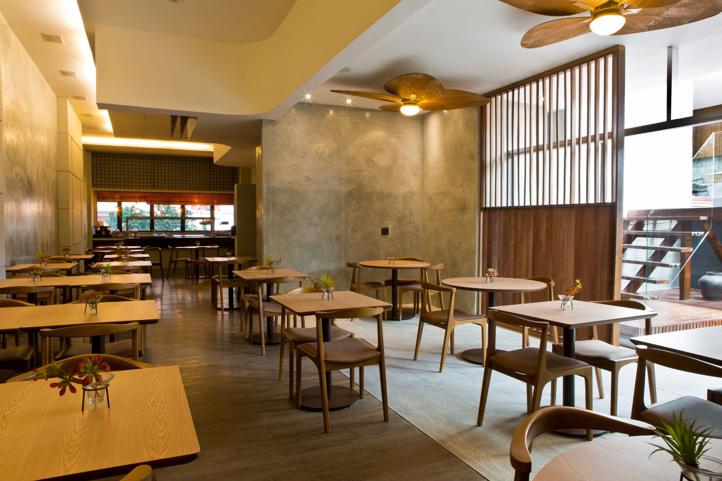 A spacious restaurant with multiple wooden tables and chairs, a ceiling fan, and large windows allowing natural light.