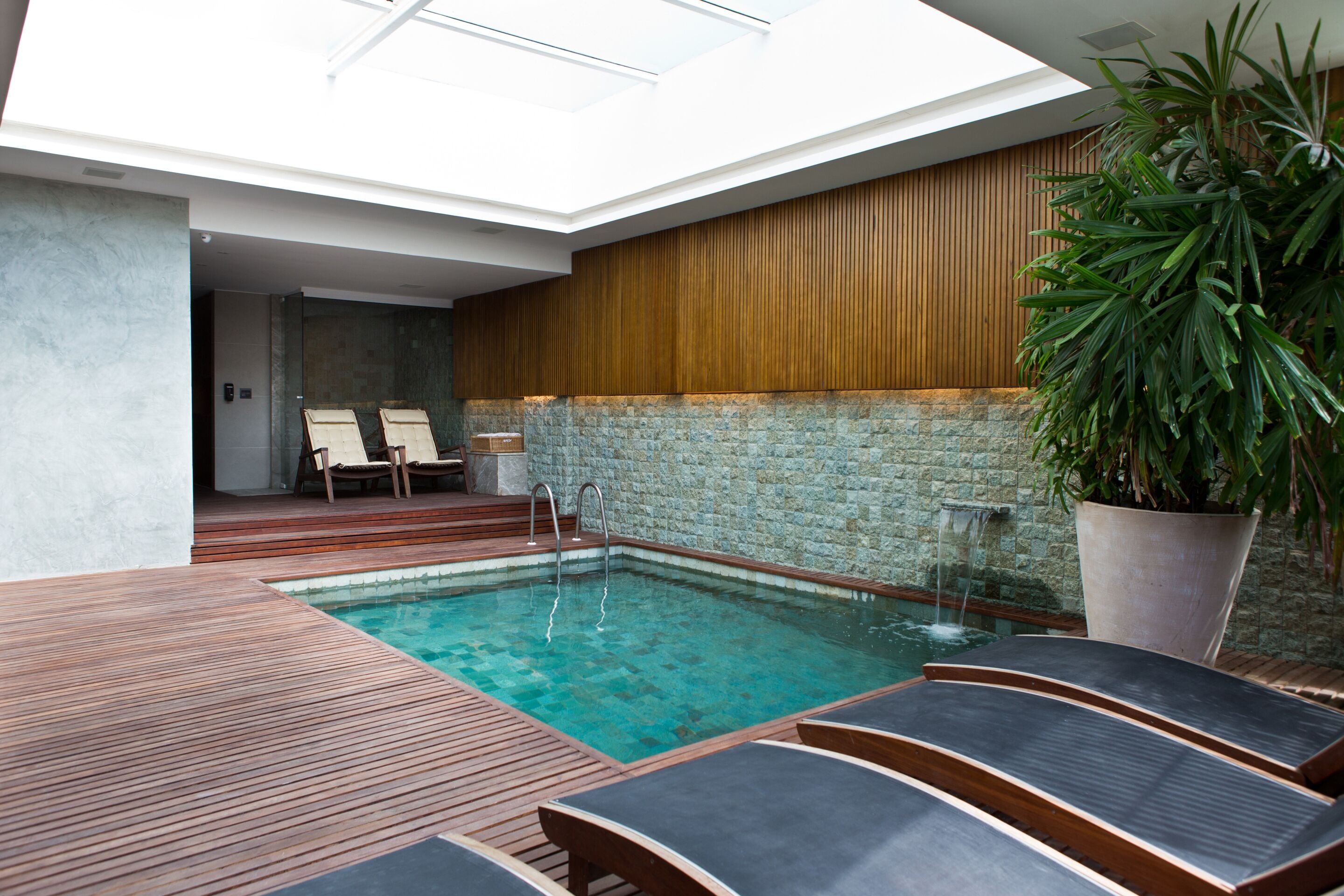 A modern indoor pool area with a wooden deck, lounge chairs, and a large potted plant.