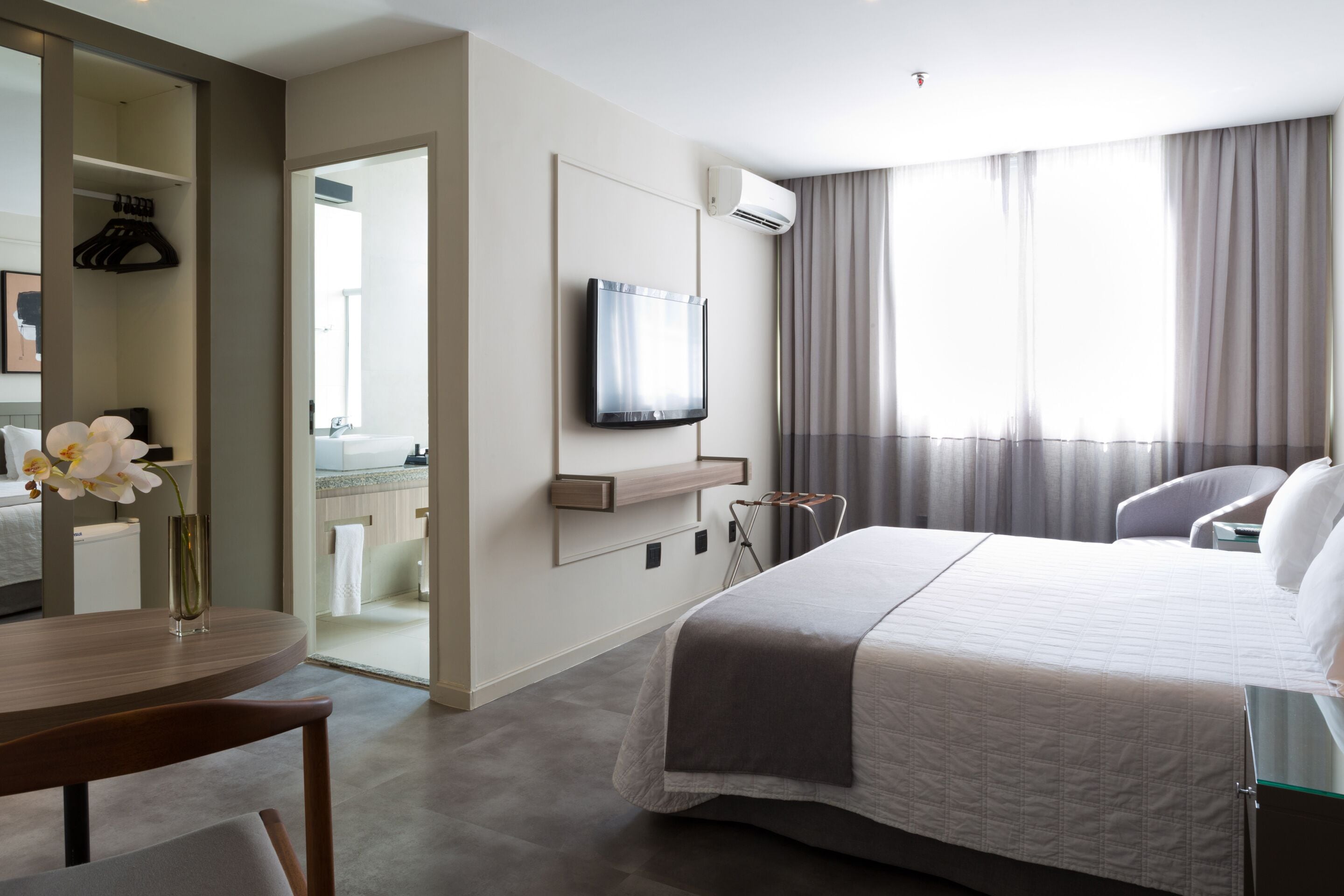 A modern hotel room with a large bed, a flat-screen TV mounted on the wall, a small table with a vase of flowers, and a bathroom visible through an open door.